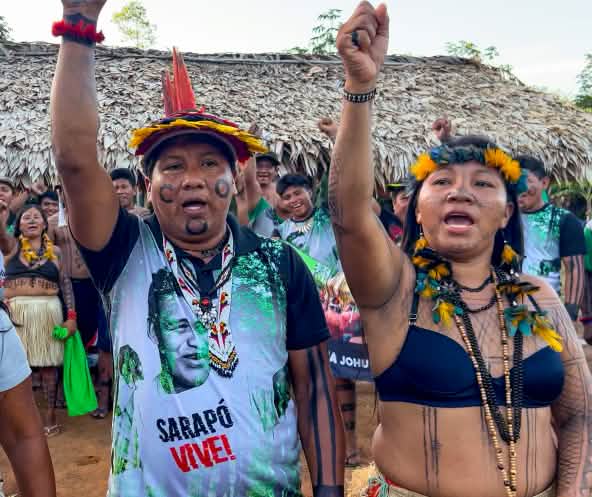 Tres indígenas Ka'apor —dos mujeres y un hombre, con los puños en alto delante de un espacio cubierto con hojas de palma.