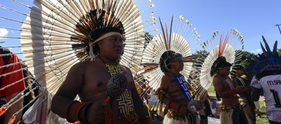 Tres hombres indígenas adornados con grandes tocados de plumas en la cabeza, sostienen unas maracas