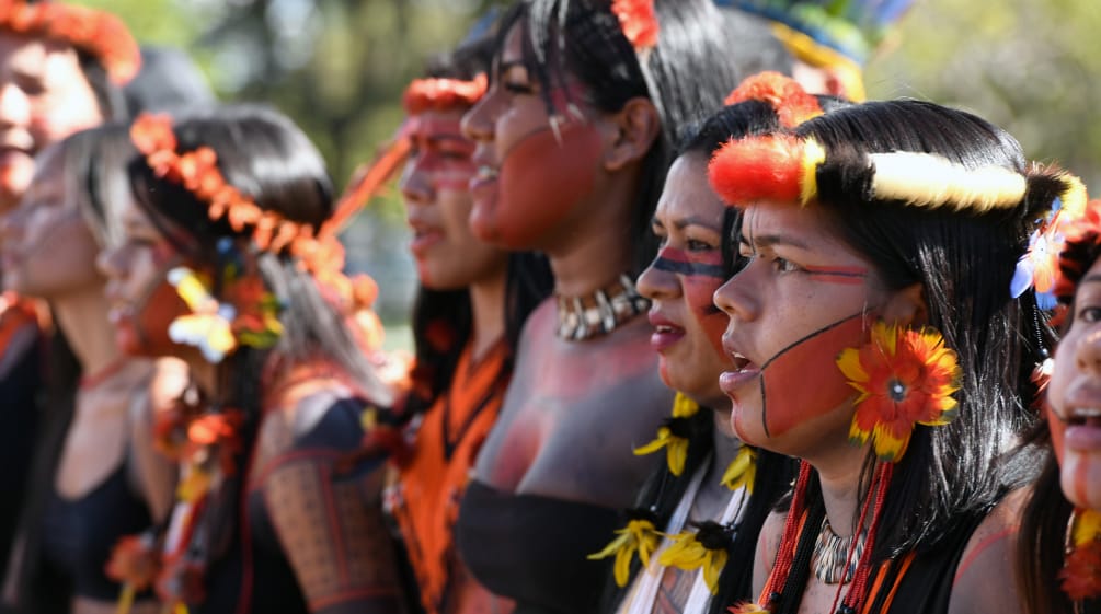 Ocho mujeres indígenas, con adornos de plumas en la cabeza, pendientes de plumas amarillas y rojas, y la cara pintada de rojo y negro