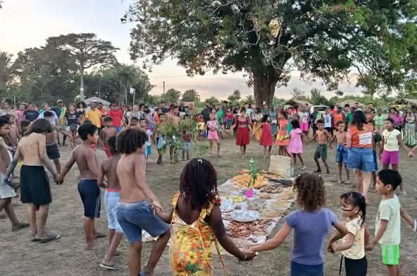 Decenas de personas, entre mujeres, hombres, niños y ancianos, forman un círculo alrededor de una ofrenda extendida en el suelo con alimentos, semillas y otros productos de la tierra (ändern).