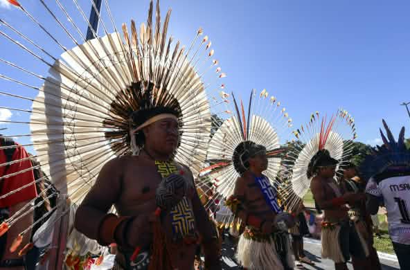 Tres hombres indígenas adornados con grandes tocados de plumas en la cabeza, sostienen unas maracas