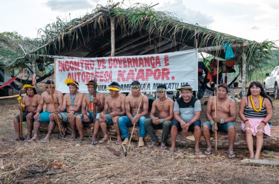 El pueblo Ka’apor en reunión en la selva amazónica. Nueve hombres y una mujer sentados en un banco ante una pancarta que dice "Encuentro de gobernanza y autodefensa Ka'apor"