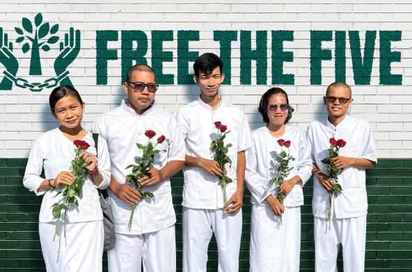Cinco personas vestidas de blanco sostienen en sus manos rosas rojas, frente a un muro en el que se lee en inglés "Liberen a los cinco" (Free the five) junto a un logo con dos manos encadenadas en medio de las cuales hay un árbol