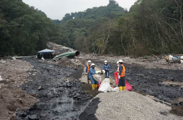 Un grupo de operadores petroleros trabaja entre dos ríos negros de petróleo que fluyen por la ladera de una montaña cubierta de rocas en la selva tropical. Al fondo se ve un tramo de tubería del oleoducto.