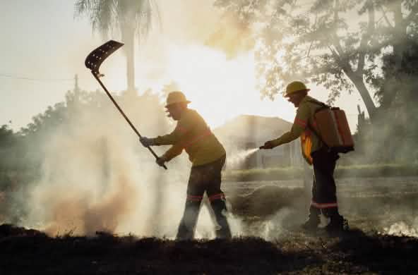 Entre humo, un hombre combate el fuego con un batefuego mientras otro hombre porta una mochila de agua y una manguera manual