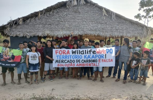 Más de 30 indígenas Ka'apor de pie frente a una casa tradicional con pilares de madera y techo de paja. Las personas sostienen una pancarta de protesta en la que se lee: "¡Fuera Wildlife Works del territorio ka'apor! El mercado de carbono es una falsa sol