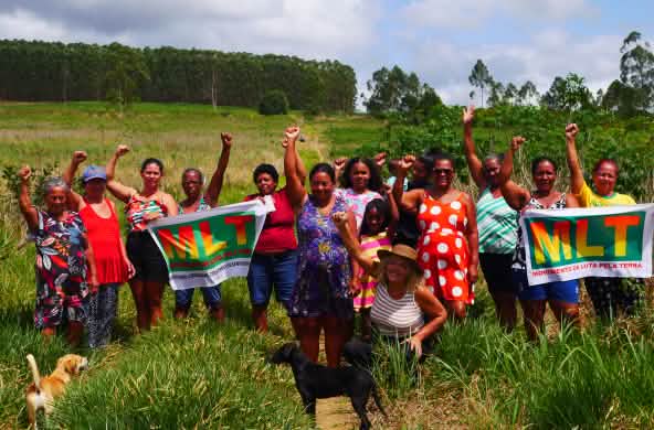 Un grupo de 15 mujeres protesta con los puños en alto y dos pancartas en las que se lee MLT Movimento de Luta pela Terra