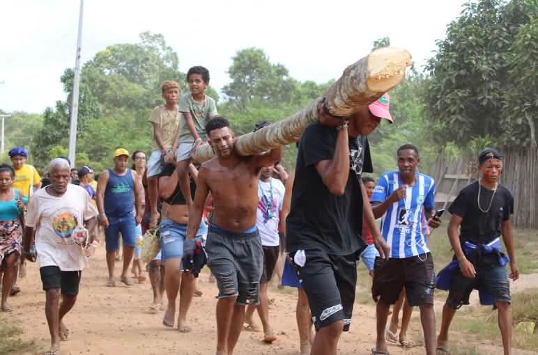 Hombres y mujeres cargan un tronco de madera en sus hombros. Dos niños van sentados sobre el tronco.