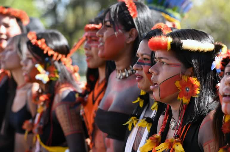 Ocho mujeres indígenas, con adornos de plumas en la cabeza, pendientes de plumas amarillas y rojas, y la cara pintada de rojo y negro