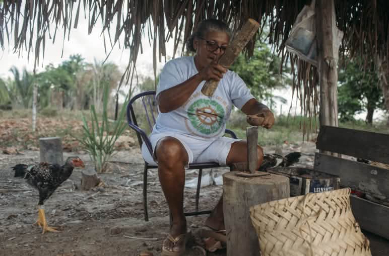 Una mujer vestida con camiseta y bermuda blanca, sentada, está rompiendo la cáscara del coco babasú utilizando dos palos a modo de herramientas