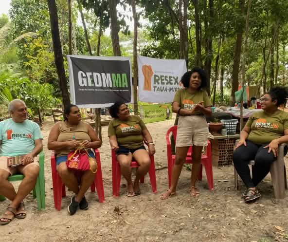 Una mujer rodeada de cuatro personas conversa a la sombra de los árboles, ante las pancartas de las tres organizaciones presentes