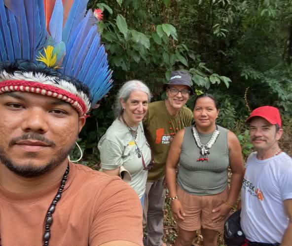 Selfie de un indígena con corona de plumas, situado a la izquierda de la imagen, junto a tres miembros de Salva la Selva y la cacica indígena Miriam Tembé. Se encuentran ante la vegetación de la selva en el estado amazónico de Pará.