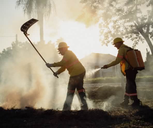 Entre humo, un hombre combate el fuego con un batefuego mientras otro hombre porta una mochila de agua y una manguera manual