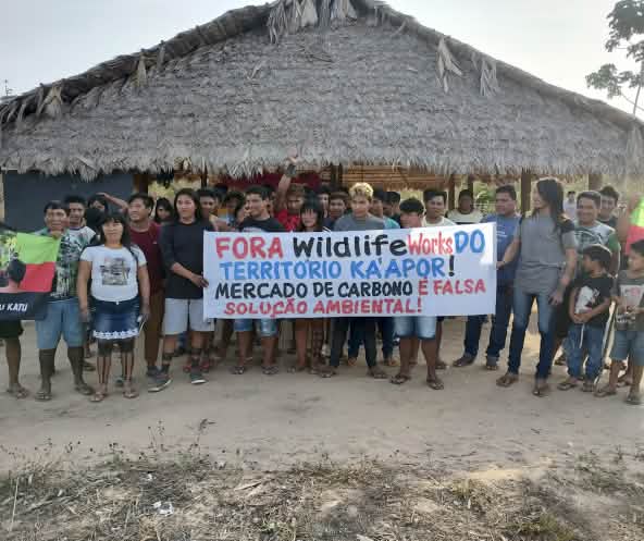 Más de 30 indígenas Ka'apor de pie frente a una casa tradicional con pilares de madera y techo de paja. Las personas sostienen una pancarta de protesta en la que se lee: "¡Fuera Wildlife Works del territorio ka'apor! El mercado de carbono es una falsa sol
