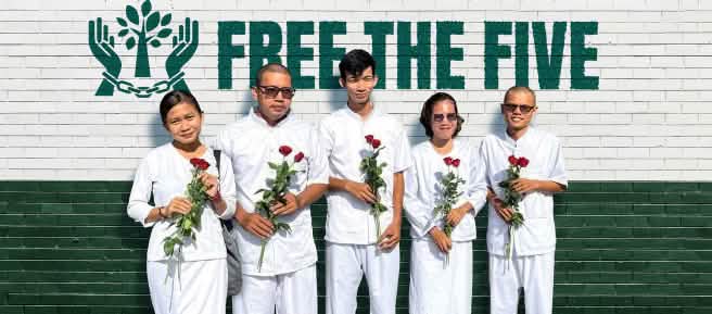 Cinco personas vestidas de blanco sostienen en sus manos rosas rojas, frente a un muro en el que se lee en inglés "Liberen a los cinco" (Free the five) junto a un logo con dos manos encadenadas en medio de las cuales hay un árbol