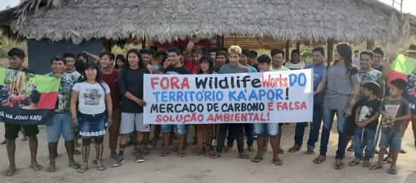 Más de 30 indígenas Ka'apor de pie frente a una casa tradicional con pilares de madera y techo de paja. Las personas sostienen una pancarta de protesta en la que se lee: "¡Fuera Wildlife Works del territorio ka'apor! El mercado de carbono es una falsa sol