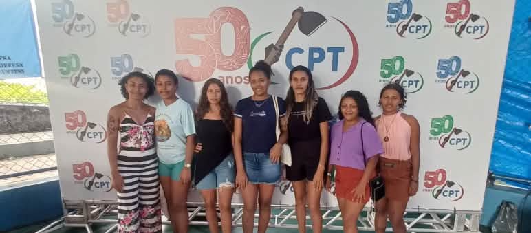 Siete mujeres jóvenes de frente y sonriendo. Detrás de ellas hay una pancarta con el logotipo 50 años de CPT (Comisión Pastoral da Terra)