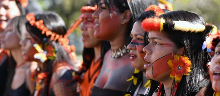 Ocho mujeres indígenas, con adornos de plumas en la cabeza, pendientes de plumas amarillas y rojas, y la cara pintada de rojo y negro