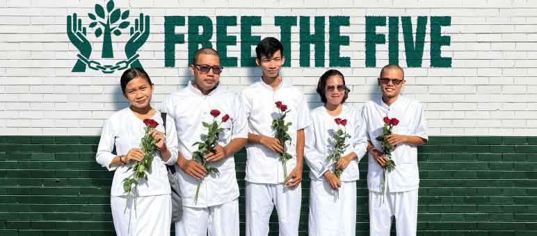 Cinco personas vestidas de blanco sostienen en sus manos rosas rojas, frente a un muro en el que se lee en inglés "Liberen a los cinco" (Free the five) junto a un logo con dos manos encadenadas en medio de las cuales hay un árbol