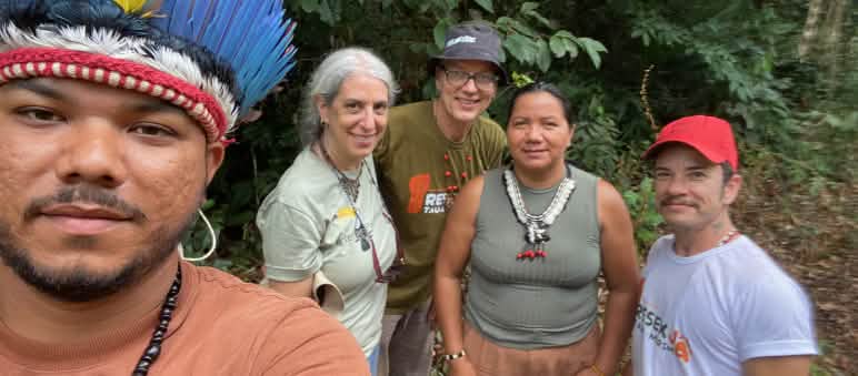 Selfie de un indígena con corona de plumas, situado a la izquierda de la imagen, junto a tres miembros de Salva la Selva y la cacica indígena Miriam Tembé. Se encuentran ante la vegetación de la selva en el estado amazónico de Pará.