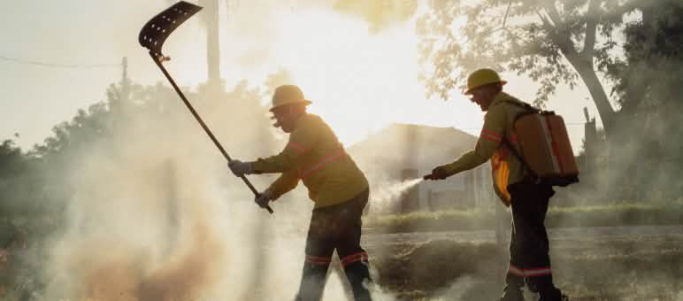 Entre humo, un hombre combate el fuego con un batefuego mientras otro hombre porta una mochila de agua y una manguera manual