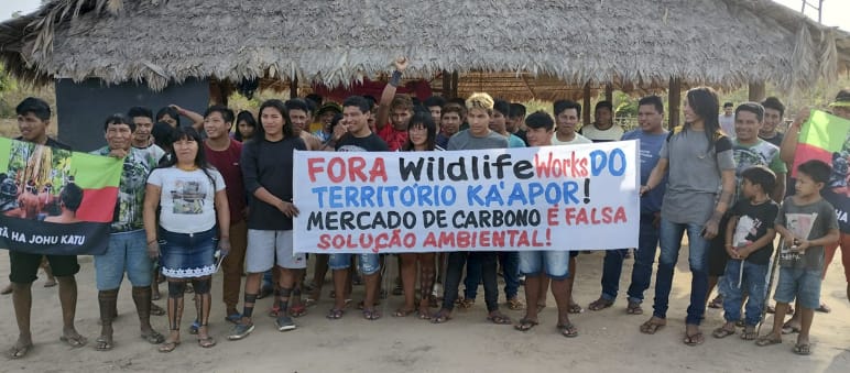 Más de 30 indígenas Ka'apor de pie frente a una casa tradicional con pilares de madera y techo de paja. Las personas sostienen una pancarta de protesta en la que se lee: "¡Fuera Wildlife Works del territorio ka'apor! El mercado de carbono es una falsa sol