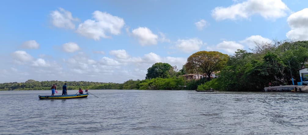 Bajo un cielo azul,  tres pescadores en una pequeña embarcación navegan mar adentro. A la derecha, se ven unas pequeñas casas en la costa, entre la vegetación.
