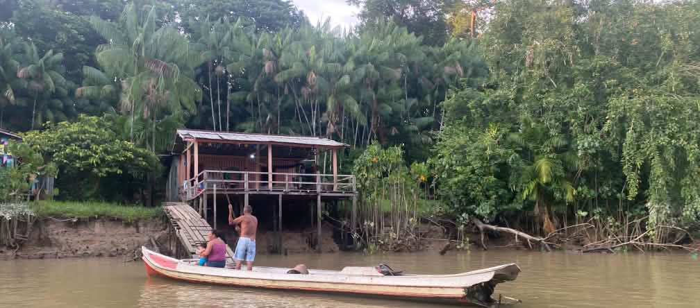 Dos personas en una canoa de madera frente a una cabaña también de madera a orillas de un río en la selva tropical