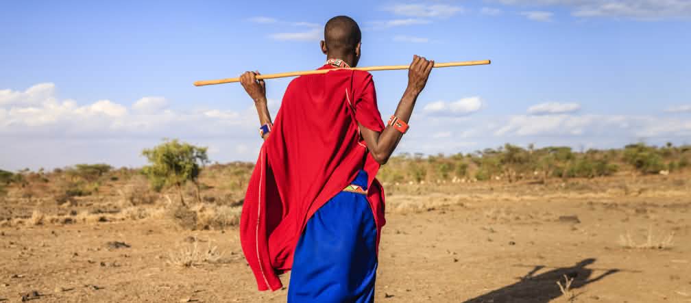 Los masái son uno de los pueblos más conocidos de África oriental Persona masái de espaldas, sosteniendo una vara de madera. La parte superior de su vestimenta es roja y la inferior, azul intenso. Su sombra se proyecta sobre el suelo.