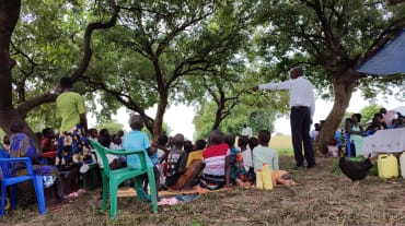 Un miembro de la organización TASHA conduce una conversación al aire libre en una comunidad, sobre el peligro de la explotación petrolífera. Está rodeado de miembros de la comunidad.