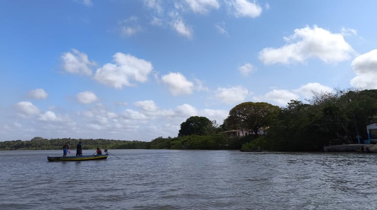 Bajo un cielo azul,  tres pescadores en una pequeña embarcación navegan mar adentro. A la derecha, se ven unas pequeñas casas en la costa, entre la vegetación.