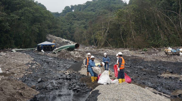 Un grupo de operadores petroleros trabaja entre dos ríos negros de petróleo que fluyen por la ladera de una montaña cubierta de rocas en la selva tropical. Al fondo se ve un tramo de tubería del oleoducto.