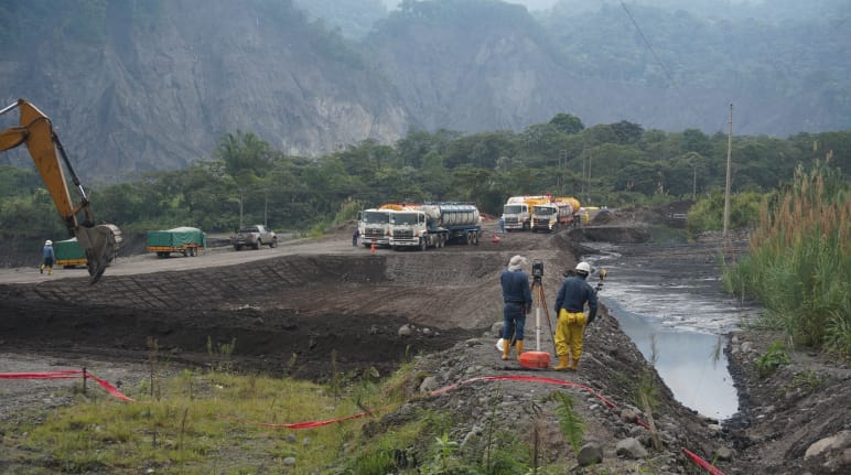 Ingenieros inspeccionan el terreno entre un estanque de tierra lleno de petróleo crudo a la derecha y una excavadora visible a la izquierda, está cavando otro pozo de tierra. Al fondo, delante de la montaña se ven camiones cisterna.