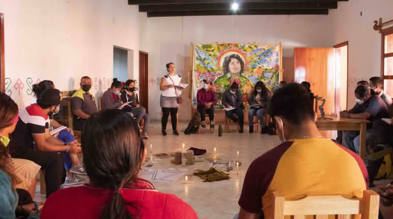 Escuela de Justicia Energética de Chiapas, pensando alternativas de autonomía en Puebla, México Personas sentadas en círculo participan de un taller, en el centro hay velas encendidas y un cartel en el que se lee la palabra PAZ y colgado en la pared un retrato dibujado de Berta Cáceres