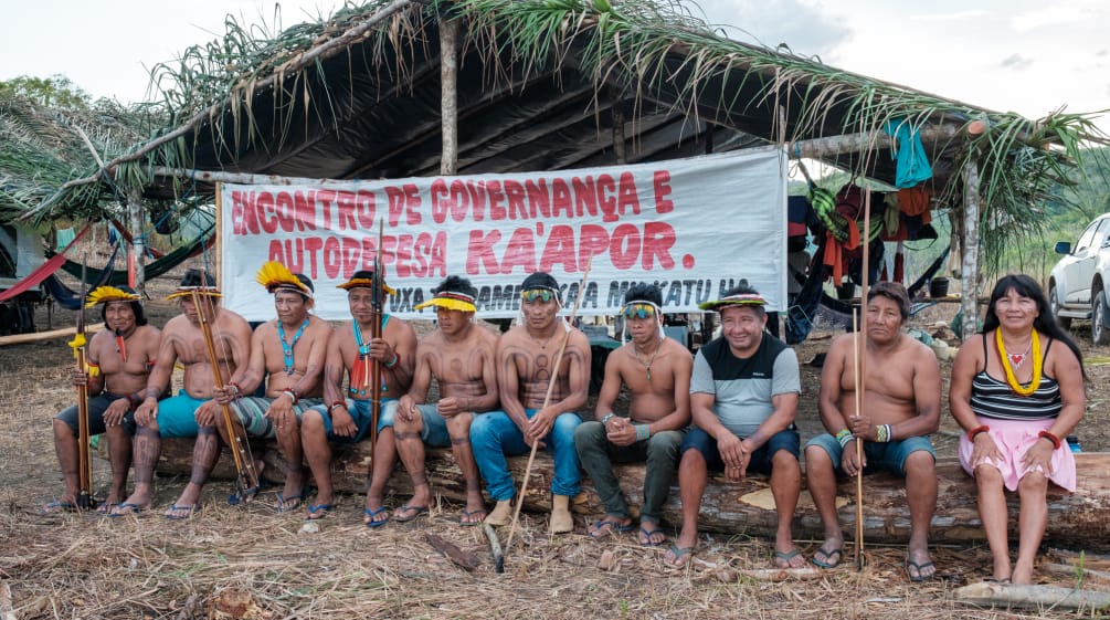 El pueblo Ka’apor en reunión en la selva amazónica. Nueve hombres y una mujer sentados en un banco ante una pancarta que dice "Encuentro de gobernanza y autodefensa Ka'apor"
