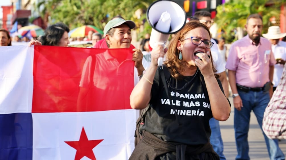 Una mujer se manifiesta en contra de la minería de cobre en Panamá, con una camiseta con la leyenda "#Panamá vale más sin minería". Al fondo la bandera panameña.