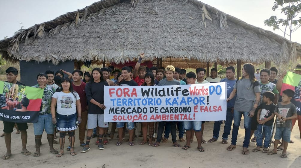 Más de 30 indígenas Ka'apor de pie frente a una casa tradicional con pilares de madera y techo de paja. Las personas sostienen una pancarta de protesta en la que se lee: "¡Fuera Wildlife Works del territorio ka'apor! El mercado de carbono es una falsa sol